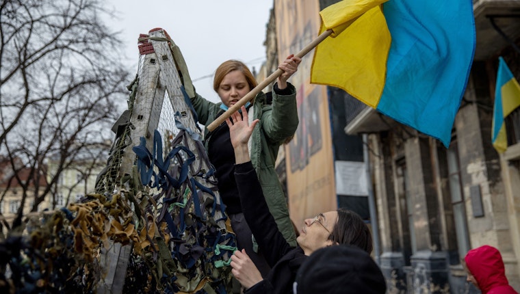 Volunteers raise a Ukrainian flag while making camouflage nets in Lviv, Ukraine, on March 1, 2022. © Ethan Swope/Bloomberg/Getty Two people lifting a Ukranian flag